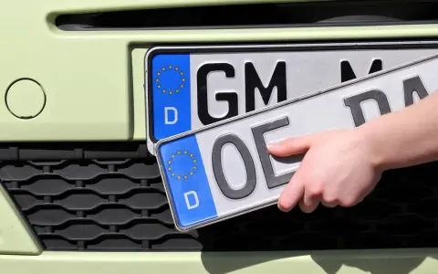 Dłoń trzyma nową tablicę rejestracyjną z literami "OE", gotową do zamontowania na samochodzie.