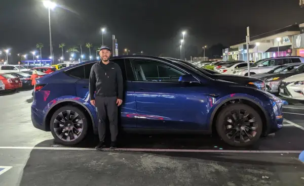Mężczyzna stoi obok ciemnoniebieskiego Tesli Model Y na nocnym parkingu.