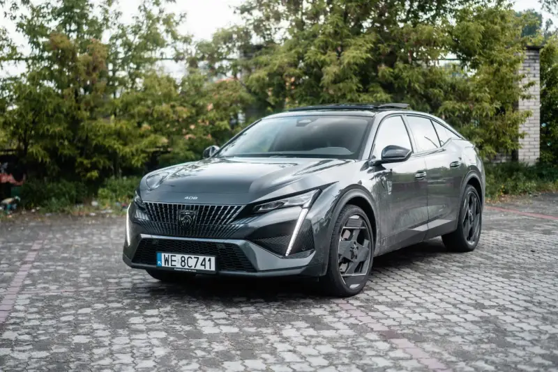 Nowoczesny Peugeot 408 Hybrid w ciemnoszarym kolorze, zaparkowany na brukowanym podjeździe, otoczony zielenią.