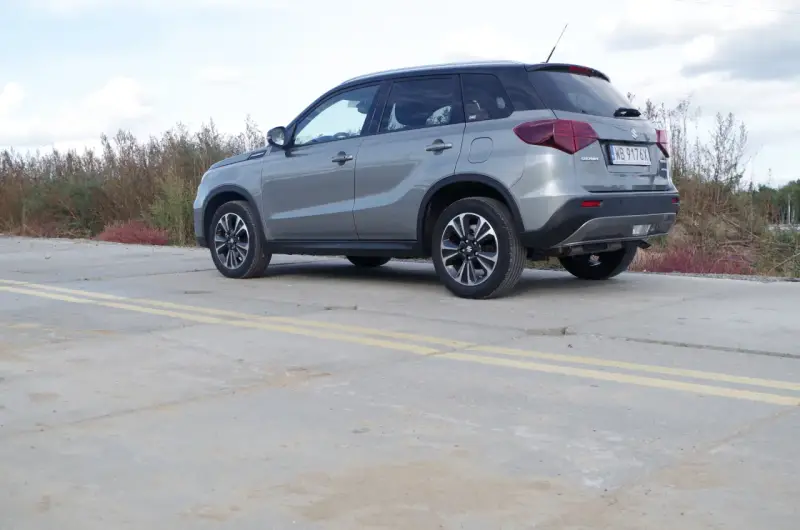 Szary Suzuki Vitara Hybrid z czarnym dachem stoi na betonowej nawierzchni z żółtymi pasami.